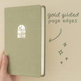 Close up of Komorebi bullet journal with green velvet cover and gold foil illustration of a window with the sun and plants