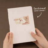 Hands holding the Junk Journal notebook with white text “textured card cover”