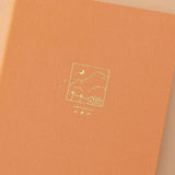 Close up of Tsuki Golden Hour bullet journal design which is an illustration of a house and some clouds in a box frame with a playlist bar underneath in gold foil on orange linen