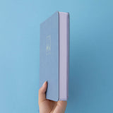 A hand holding a blue leather notebook showing the white page edges