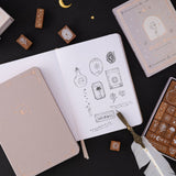 Tsuki ‘Moonlit Alchemy’ stamps on white page bullet journal and a feather quill on the side