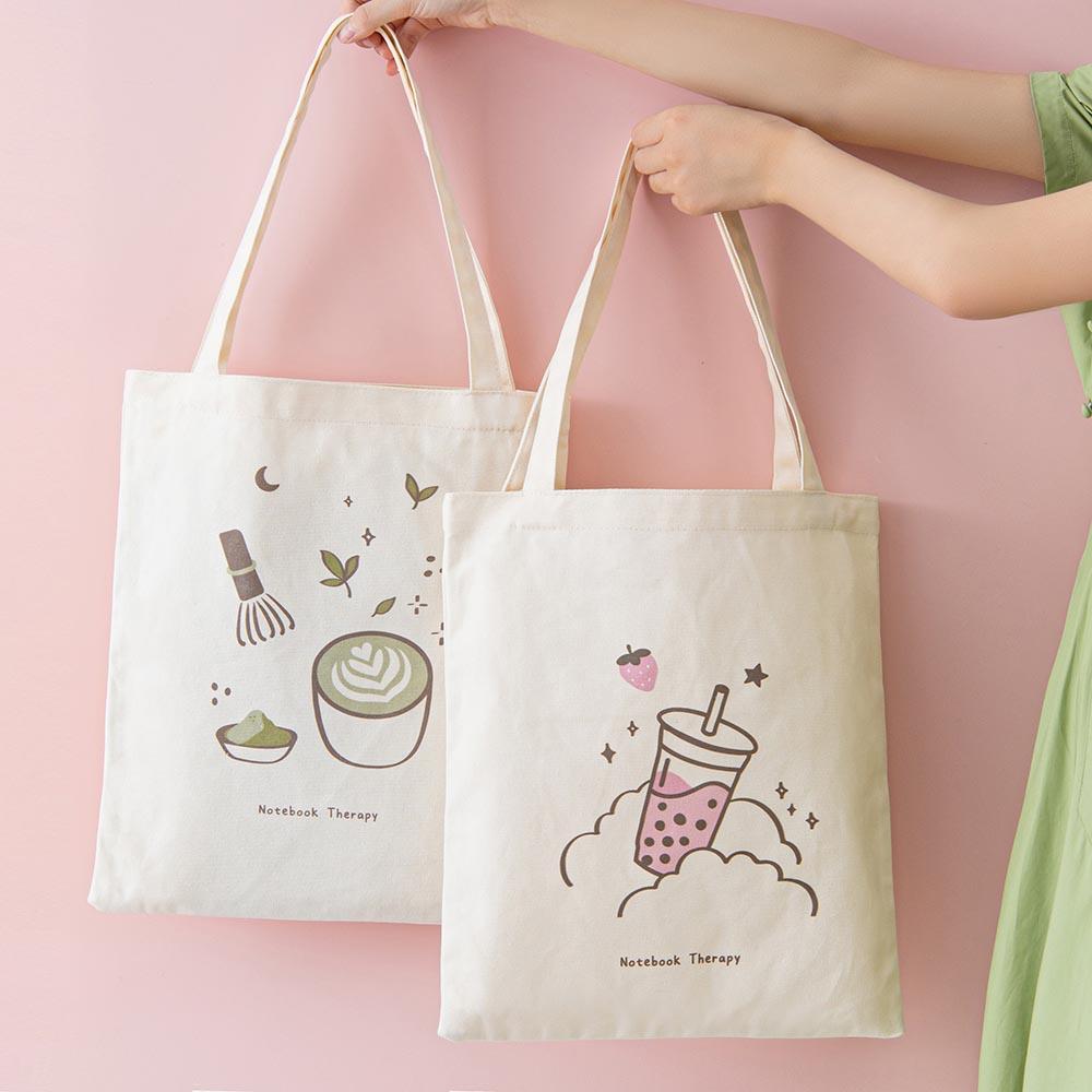 Tsuki ‘Ichigo’ Boba Tote Bag held in hands with Tsuki ‘Matcha Matcha’ Tote Bag in light pink background