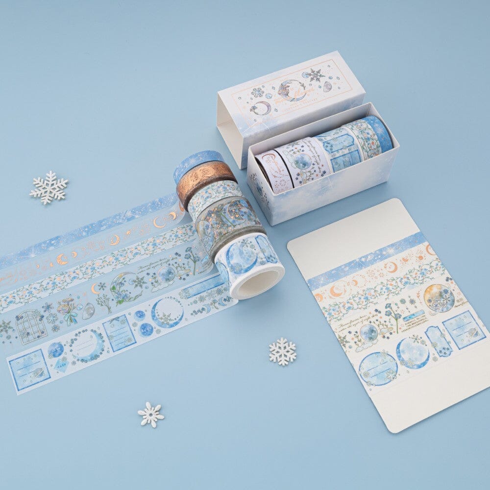 Flatlay of Winter Moonflower washi tapes with opened box and 5 washi tapes unrolled on blue background
