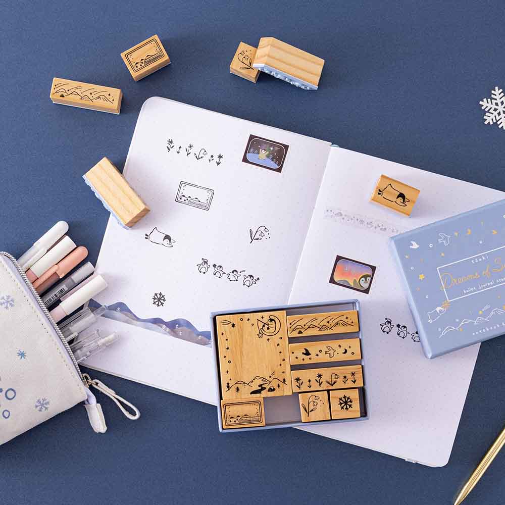 Tsuki ‘Dreams of Snow’ Bullet Journal Stamp Set and Tsuki ‘Dreams of Snow’ Holographic Washi Tapes on open bullet journal spread with Tsuki ‘Dreams of Snow’ Pop-Up Pencil Case in Playful Penguin with snowflake on navy background