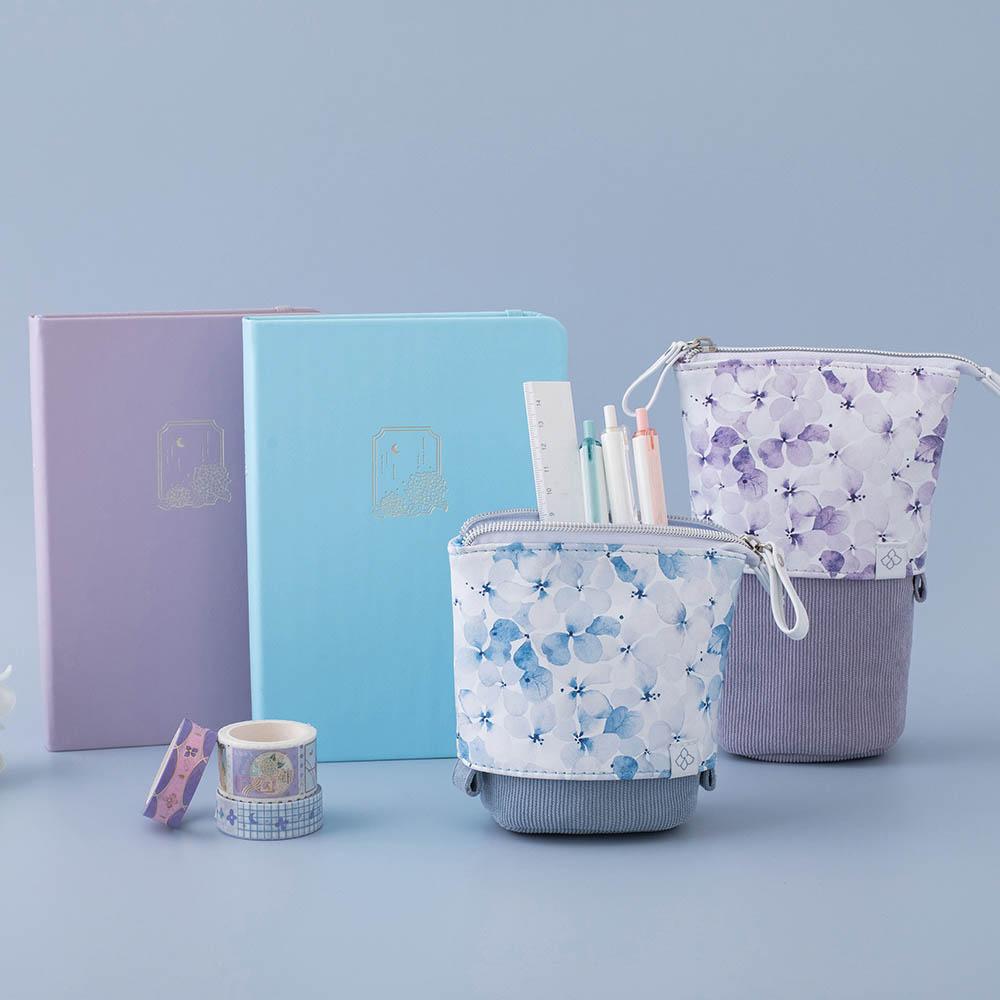 Tsuki Endless Summer Pop-Up Pencil cases in Lilac Bloom and Petal Blue with Tsuki Endless Summer Washi Tape rolls and Tsuki Endless Summer Limited Edition Bullet Journals in light blue backgroundTsuki Endless Summer Pop-Up Pencil cases in Lilac Bloom and Petal Blue on stacked Tsuki Endless Summer Limited Edition Bullet Journals with Tsuki Endless Summer Washi Tape rolls in eco-friendly gift box packaging in light blue background