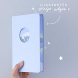 Tsuki Four Seasons Winter Collector’s Edition Bullet Journal held in hands at an angle in light blue background