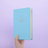 Tsuki Endless Summer Limited Edition Bullet Journal in Petal Blue held in hands at spine angle in lilac background