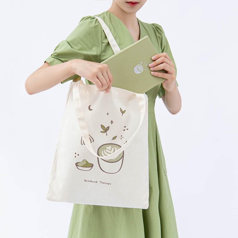 Tsuki ‘Matcha Matcha’ Tote Bag shown carried by model with Tsuki ‘Matcha Matcha’ Limited Edition Bullet Journal in light grey background