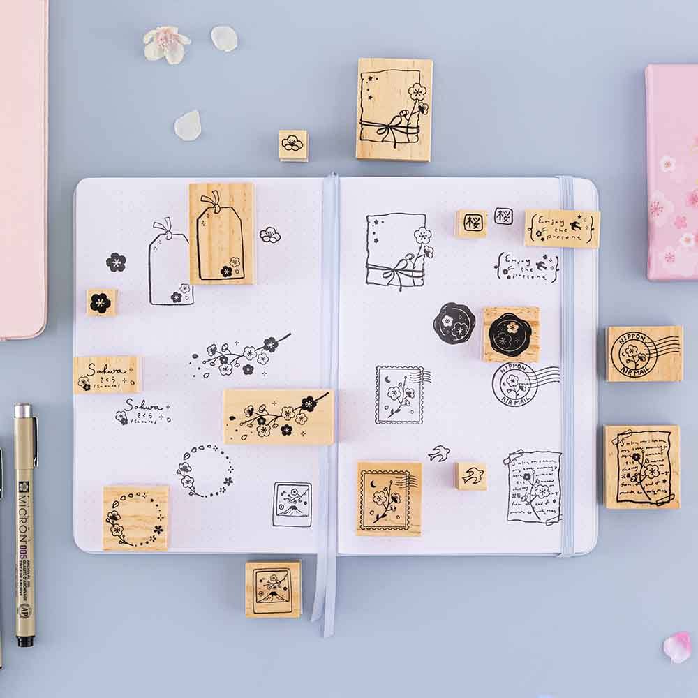 Tsuki ‘Sakura Journey’ Bullet Journal Stamps on open Tsuki ‘Sakura Journey’ Limited Edition Bullet Journal with pen and cherry blossom petals on light blue background