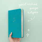 Tsuki Teal Sky ‘Flutter + Dream’ Limited Edition Bullet Journal by Notebook Therapy x Pelinkan with gold etched page edges held in hand at an angle in mint background