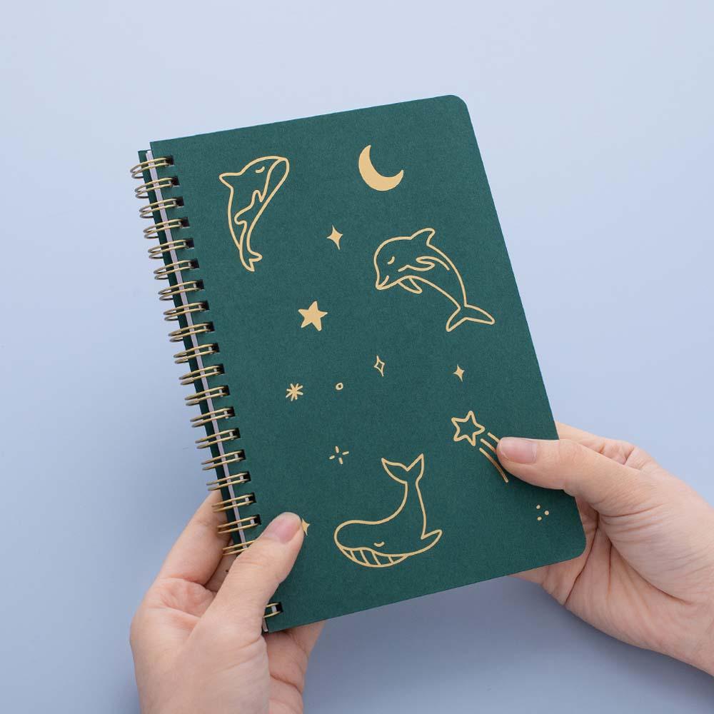 Tsuki Ocean Edition Ring Bound notebook in deep teal held in hands in blue background