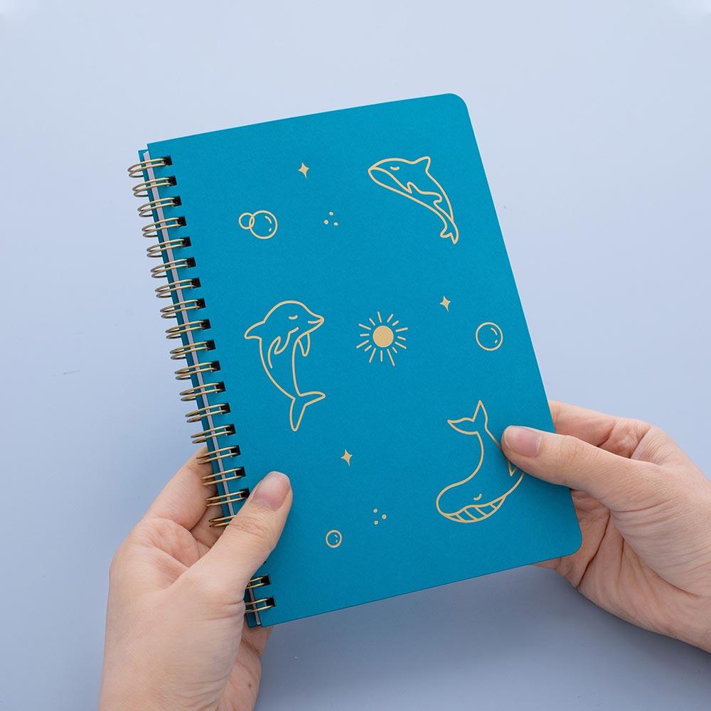 Tsuki Ocean Edition Ring Bound notebook in aqua blue held in hands in blue background