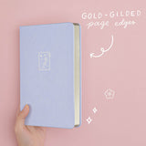 Tsuki ‘Sakura Journey’ Limited Edition Bullet Journal held in hand at spine angle with gold gilded page edges in pink background