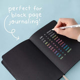 Gelly Roll Pen in white held in hand over open Tsuki Black Paper Limited Edition Hardcover Bullet Journal on light teal background