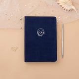 Tsuki deep blue textured vegan leather Gentle Giant luxury edition notebook with gold pen and seashells on peach background