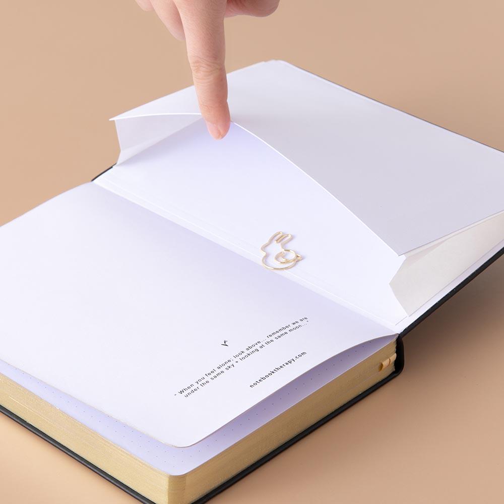 Close up of the open back page pocket of Tsuki ‘Moonlit Wish’ Limited Edition Bullet Journal held in hand with free gift on light brown background