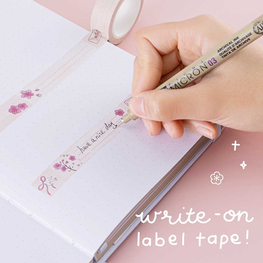 Close up of Tsuki ‘Sakura Journey’ Writing Washi Tape on open bullet journal page with pen held in hand on light pink background