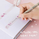 Close up of Tsuki ‘Sakura Journey’ Writing Washi Tape on open bullet journal page with pen held in hand on light pink background