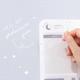 Close up of the front of Tsuki Reusable Weekly Planner with protective film being removed on light grey surface