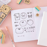 Close up of Tsuki ‘Flutter + Dream’ Bullet Journal Stamp Set by Notebook Therapy x Pelinkan with frame stamps on open Tsuki Teal Sky ‘Flutter + Dream’ Limited Edition Bullet Journal by Notebook Therapy x Pelinkan with Tsuki Cloud White White Sky ‘Flutter + Dream’ Limited Edition Bullet Journal by Notebook Therapy x Pelinkan with gelly pen on pastel pink background