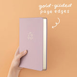 Tsuki Blush Pink ‘Bunny Blush’ Limited Edition Bullet Journal designed with Blossom Bujo with gold gilded page edges held in hands in lilac background