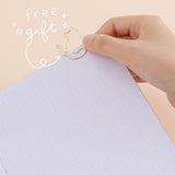 Hand putting cat-shaped paperclip in gold on a bullet journal page with the words ‘free gift’ in white lettering