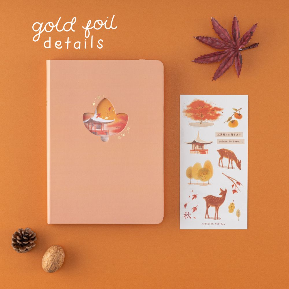 Orange bullet journal with a maple leaf cutout and text that says ‘gold foil details’ in white and a sticker sheet with autumn illustrations on it