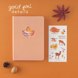 Orange bullet journal with a maple leaf cutout and text that says ‘gold foil details’ in white and a sticker sheet with autumn illustrations on it