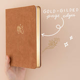 Hand holding up brown velvet bullet journal with gold gilded page edges