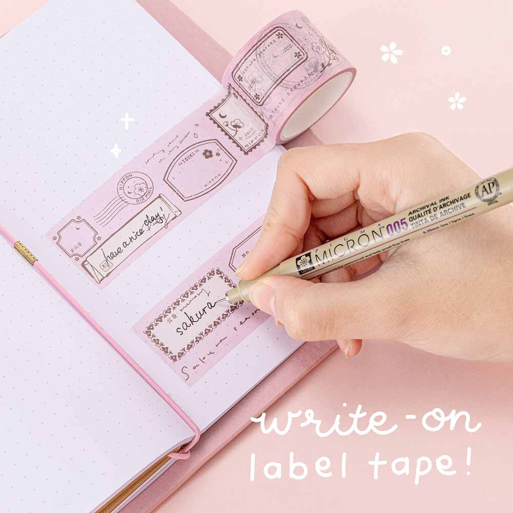 Close up of Tsuki ‘Sakura Journey’ Vintage Journal Write on label tape with micron pen held in hand over Tsuki ‘Sakura Journey’ Limited Edition Travel Notebook on pink background