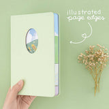 Hand holding Tsuki Four Seasons Summer Collectors Edition 2022 sage bullet journal notebook on sage green background with dried flower decoration and “illustrated page edges” in white handwritten text